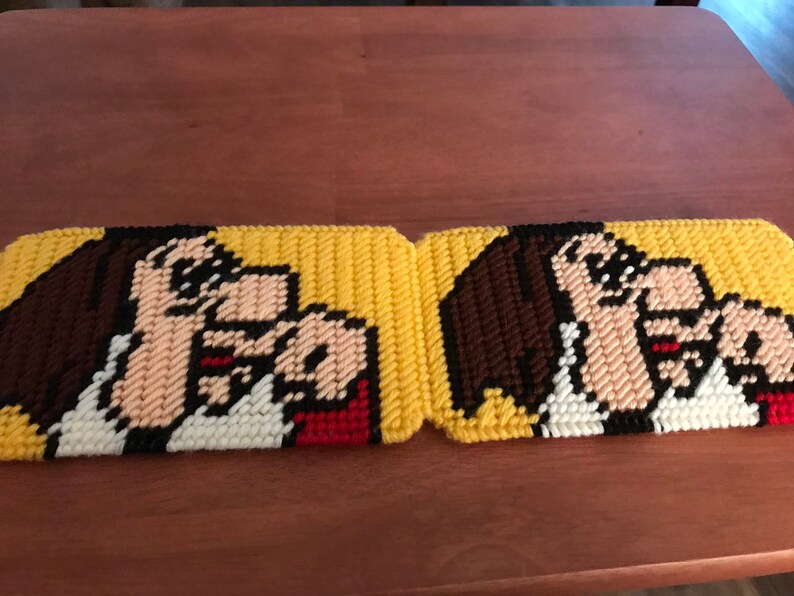May include: Two yellow and brown crocheted coasters with a cartoon image of a woman with brown hair and a red and white shirt. The coasters are made of plastic canvas.