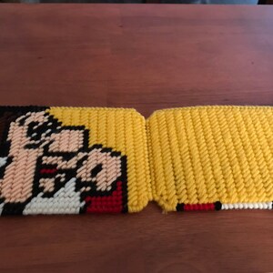 May include: Two yellow and white crocheted pot holders with a cartoon image of a person with brown hair and a red mouth. The person is pointing with their right hand.