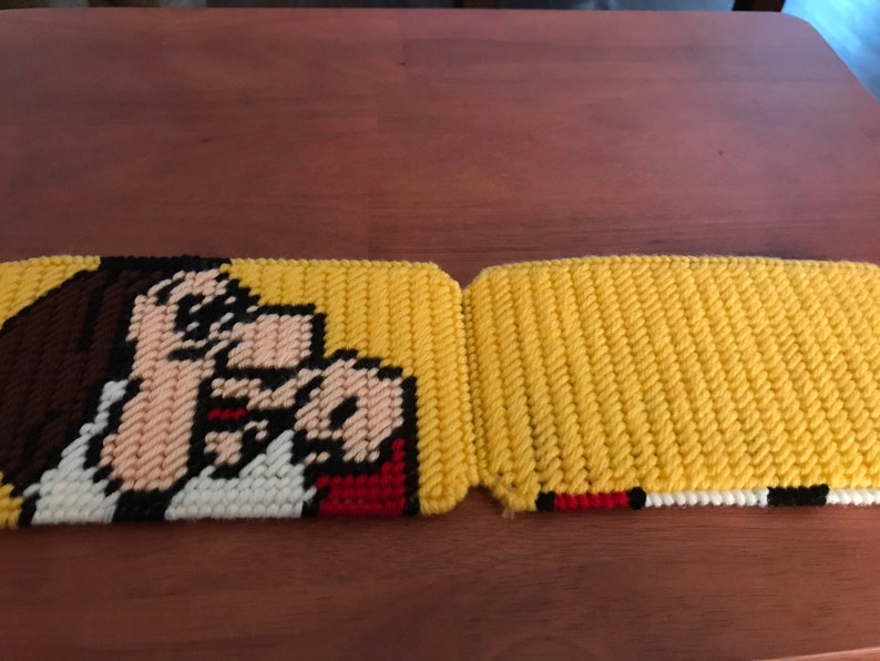 May include: Two yellow and white crocheted pot holders with a cartoon image of a person with brown hair and a red mouth. The person is pointing with their right hand.
