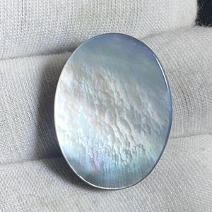 May include: Oval-shaped iridescent gemstone with a pearlescent sheen. The surface displays a cloudy, textured pattern with hints of blue, white, and subtle rainbow hues. The gemstone is set against a white fabric background.