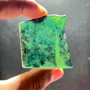 Può includere: Una gemma rettangolare e traslucida con una vibrante tonalità verde, punteggiata da inclusioni verdi e nere più scure. La pietra è tenuta tra le dita, mostrando i suoi bordi naturali e irregolari e la superficie lucida. La gemma è un pezzo unico di arte naturale.