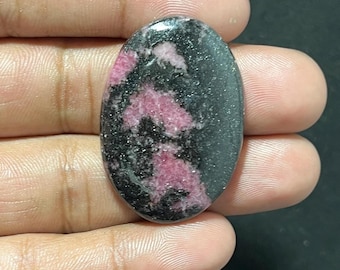 Puede incluir: Gema ovalada pulida, con una mezcla de negro y rosa. El rosa aparece en un patrón moteado. La gema se sostiene en una mano, mostrando su tamaño y forma.