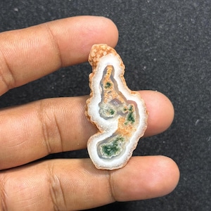 Puede incluir: Una piedra pulida de forma irregular con un patrón blanco, verde y naranja. La piedra tiene un borde exterior blanco, una banda central blanca e inclusiones verdes y naranjas. La piedra se sostiene en una mano.