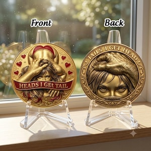 May include: Two circular gold-toned metal coins displayed on clear plastic stands. The left coin features a figure with red hearts and the text 'HEADS I GET TAIL' on a red banner. The right coin shows a hand on a face with the text 'TAILS I GET HEAD'.