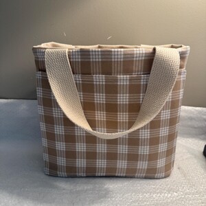 May include: A beige and white plaid tote bag with a cream-colored handle. The bag has a rectangular shape and a pocket on the front. The interior of the bag is a solid cream color.