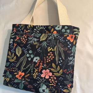 May include: A tote bag with a floral design. The bag has a dark background adorned with orange, pink, blue, and green flowers and leaves. It has a cream-coloured handle.
