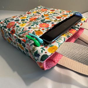 May include: A floral print tote bag with a tablet inside. The bag features a white background with red, yellow, and pink flowers and green leaves. The interior is pink with white hearts. Two pens are visible.