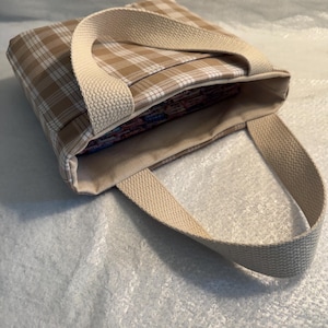 May include: A beige and white checked tote bag with cream-coloured handles. The bag is open, revealing the contents inside. The bag is made of a sturdy fabric and is designed for carrying items.