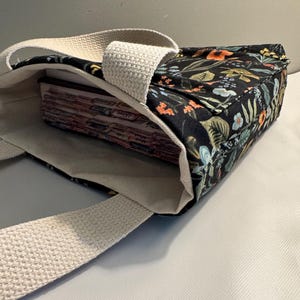 Puede incluir: Bolso tote negro con estampado floral en naranja, verde y azul. El bolso tiene forro y correas de lona color crema. El bolso está abierto, revelando su contenido. Adecuado para llevar libros u otros objetos.