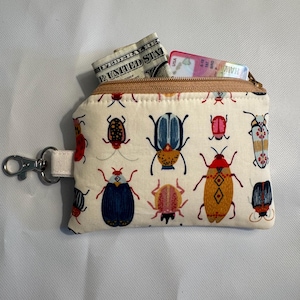 May include: A small, cream-colored zippered pouch with a repeating pattern of colorful beetles. The pouch is open, revealing a folded US dollar bill and a credit card. A silver-colored clasp is attached to the side.