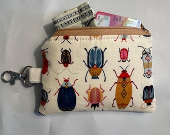 May include: A small, cream-coloured zippered pouch featuring a repeating pattern of colourful beetles. The pouch is open, showing a folded banknote and a credit card. A silver-coloured clasp is attached to the side.
