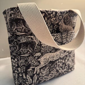 May include: A small tote bag with a cream-colored strap. The bag is black with a white forest print featuring animals and foliage. The strap is made of a woven material.