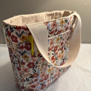 Puede incluir: Un bolso tote con estampado floral con asa y forro interior color crema. El bolso presenta un colorido estampado de flores rojas, amarillas y rosas sobre un fondo blanco. Una cremallera amarilla es visible en el bolsillo delantero.