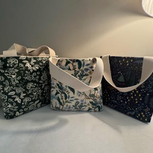May include: Three fabric tote bags with cream-colored handles. The bags feature floral and botanical patterns in shades of green, blue, and cream. The bags are designed for carrying items.