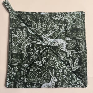 May include: Square, dark green pot holder with a white woodland design featuring rabbits, squirrels, and foliage. The pot holder has a quilted pattern and a small loop for hanging. A charming kitchen accessory.