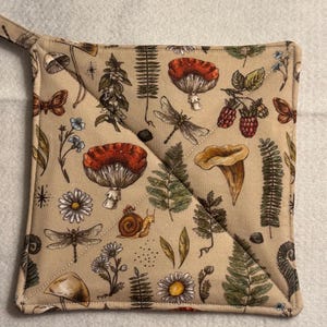 Op de afbeelding: Beige vierkante pannenlap met een ophanglus. De stof is bedrukt met een patroon van paddenstoelen, libellen, bloemen, varens en bessen in rood, groen en bruin. De pannenlap heeft een gewatteerd ontwerp.