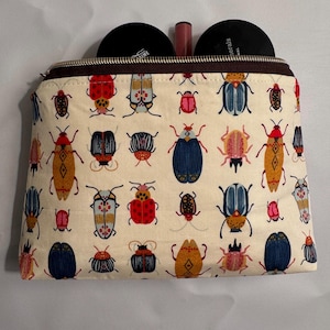 May include: Beige zippered cosmetic bag with a colorful insect pattern. The bag contains two black compacts and a tube of lipstick. The bag's zipper is brown, and the fabric is covered in various beetle illustrations.