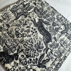 May include: Black and white quilted pot holder with a loop for hanging. The fabric features a repeating pattern of rabbits, flowers, and foliage. The design is a whimsical, nature-inspired illustration.