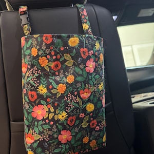May include: A floral print car organizer bag, featuring a black background with red, orange, yellow, and pink flowers and green leaves. The bag hangs from the car seat headrest with a black buckle and strap.