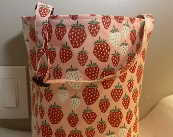 May include: A pink fabric tote bag with a repeating pattern of red and white strawberries. The bag has a drawstring closure and a handle. The strawberries have green leaves and are scattered across the pink background.