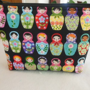 May include: Black cosmetic bag with a zip closure, patterned with colourful Matryoshka dolls. The dolls feature floral and geometric designs, with diverse skin tones and hair colours. The bag is rectangular.
