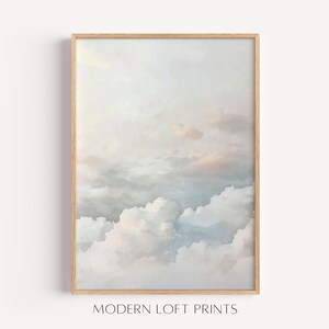 Op de afbeelding: Een ingelijste kunstdruk met een aquarel schilderij van een bewolkte lucht. Het kunstwerk toont zachte pastelkleuren, waaronder tinten blauw, wit en perzik. De print zit in een licht houten frame, en de tekst "MODERN LOFT PRINTS" staat onderaan.
