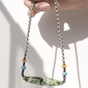 Könnte beinhalten: Eine handgefertigte Halskette mit einer länglichen Mittelperle mit einem gesprenkelten grün-braunen Muster. Die Halskette hat eine geflochtene Kordel in Grau, Schwarz und Weiß mit orangefarbenen und blauen Akzentperlen. Die Halskette wird vor einem weißen Hintergrund gehalten.