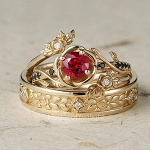 Può includere: Un set di tre anelli d'oro. L'anello di fidanzamento presenta una gemma rossa rotonda circondata da un design floreale. Le fedi nuziali hanno motivi a foglie e fiori con piccoli diamanti e gemme nere. Gli anelli sono su uno sfondo neutro.