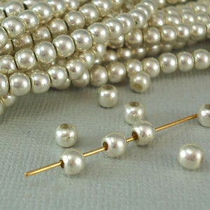 May include: A close-up of a pile of white pearl beads with a few beads strung on a gold wire. The beads are round and have a smooth, shiny finish.