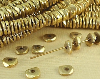 May include: A collection of metallic gold-colored beads with a flattened, irregular triangular shape. The beads feature a central hole and a textured, reflective surface. Several strands of these beads are coiled together, with loose beads scattered.