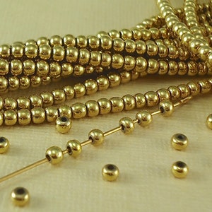May include: Close-up of gold-colored beads, strung and loose. The beads are small, round, and have a metallic sheen. Some beads are strung on a thin gold wire, while others are scattered. The image suggests jewelry making or craft supplies.