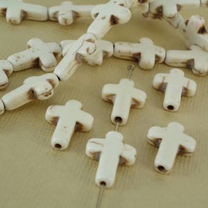 May include: Ivory-colored cross-shaped beads with brown accents are strung together, some loose and some on a string. The beads have a matte finish and are arranged on a light tan surface. The image shows a close-up of the beads.