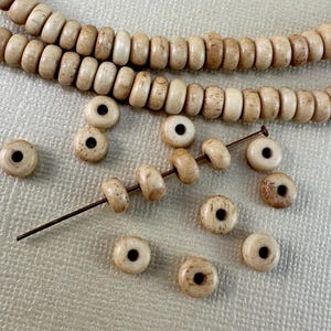 May include: A collection of round, disc-shaped beads with a natural tan color and speckled texture. One long strand of beads is arranged in a curved line, while several individual beads are scattered below, with five beads threaded onto a thin metal pin.
