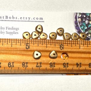 Puede incluir: Peque&ntilde;as cuentas triangulares doradas con un agujero central, mostradas con una regla de madera. La regla muestra medidas en cent&iacute;metros. Tambi&eacute;n hay una tarjeta de visita con el texto "BestBobs.etsy.com". Para hallazgos y suministros de joyer&iacute;a.