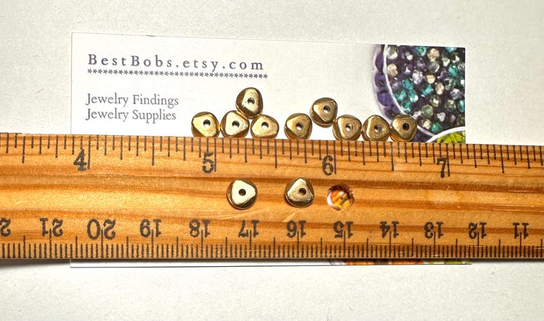 Puede incluir: Peque&ntilde;as cuentas triangulares doradas con un agujero central, mostradas con una regla de madera. La regla muestra medidas en cent&iacute;metros. Tambi&eacute;n hay una tarjeta de visita con el texto "BestBobs.etsy.com". Para hallazgos y suministros de joyer&iacute;a.