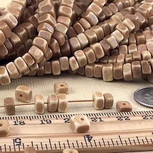 May include: A strand of light brown ceramic square beads with a slightly irregular shape. The beads are approximately 1/2 inch in size and are strung on a white cord. The beads are shown on a wooden ruler with inch markings.