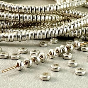 May include: Silver metal beads in a round shape with a hole in the center. The beads are arranged in a pile and on a string.
