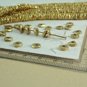 Puede incluir: Primer plano de cuentas de metal de color dorado. Las cuentas son peque&ntilde;as, planas y de forma cuadrada con un agujero en el centro. Algunas cuentas est&aacute;n ensartadas, mientras que otras est&aacute;n sueltas. Una regla est&aacute; en el marco, lo que sugiere medidas.