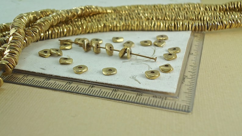 Puede incluir: Primer plano de cuentas de metal de color dorado. Las cuentas son peque&ntilde;as, planas y de forma cuadrada con un agujero en el centro. Algunas cuentas est&aacute;n ensartadas, mientras que otras est&aacute;n sueltas. Una regla est&aacute; en el marco, lo que sugiere medidas.