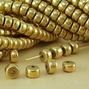May include: A close-up of gold-colored round beads. The beads are small and have a smooth, shiny finish. They are arranged in a pile and scattered on a light brown surface.