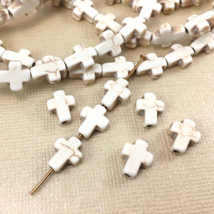May include: White cross-shaped beads with brown marbling. The beads are strung on a wire and some are loose. The beads are on a beige surface.