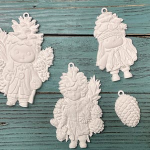 May include: Four white Christmas ornaments. Three ornaments are shaped like children in winter attire, and one is a pinecone. The ornaments have a loop at the top for hanging. The ornaments are on a teal wooden surface.