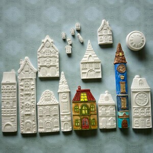 Może przedstawiać: Kolekcja miniaturowych ceramicznych domków o różnych kształtach i rozmiarach. Niektóre są białe, inne mają kolorowe detale, takie jak czerwone, żółte i niebieskie dachy i akcenty. W zestawie znajduje się również wieża zegarowa i okrągły zegar.