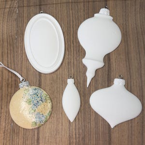 Puede incluir: Una colección de adornos navideños de varias formas. Incluye un adorno redondo con diseño floral, un marco ovalado y tres adornos blancos de diferentes formas, listos para decorar.