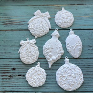 Puede incluir: Una colección de adornos navideños blancos y ornamentados en varias formas, incluyendo diseños redondos, de lágrima y ovalados. Cada adorno presenta patrones florales y de lazo detallados, con pequeños lazos para colgar. Los adornos están dispuestos sobre una superficie de madera turquesa.