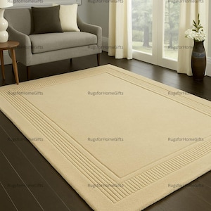 Könnte beinhalten: Beiger rechteckiger Teppich mit strukturiertem Rand und glatter Mitte. Der Teppich liegt auf einem dunklen Holzboden in einem Wohnzimmer. Der Raum umfasst ein graues Sofa, Vorhänge und eine Vase mit Blumen. Maße in Zentimetern.