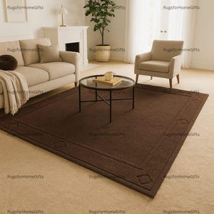 May include: A rectangular brown rug with a diamond pattern border is displayed in a living room setting. The rug is placed on a light beige carpet, with a sofa, armchair, and coffee table visible in the background. The rug measures approximately 8 x 10 feet.