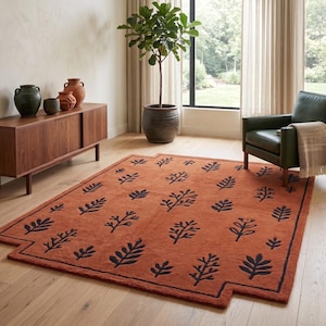 Puede incluir: Una alfombra rectangular de color naranja quemado con un patrón repetitivo de motivos de hojas botánicas en azul marino oscuro. La alfombra tiene un borde grueso y esquinas con muescas, creando una forma geométrica distintiva en el suelo.