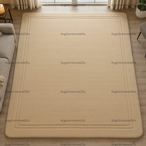 May include: A rectangular beige rug with a bordered design. The rug has a soft, plush texture and is placed on a wooden floor. The rug is a neutral colour, suitable for various interior design styles.