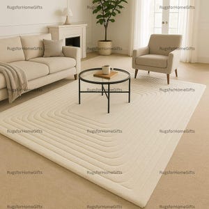 May include: A cream-colored, rectangular rug with a concentric, curved design. The rug is placed in a living room setting with a sofa, armchair, and a round coffee table. The room has a neutral color palette.